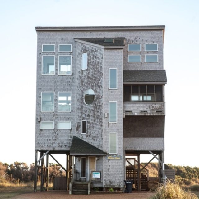 Tall, weathered, gray beach house on stilts with many windows, a small entryway, and grass in the foreground.