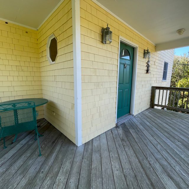 Wooden porch with yellow shingle siding, a green table and chairs, green door, and two wall lanterns.