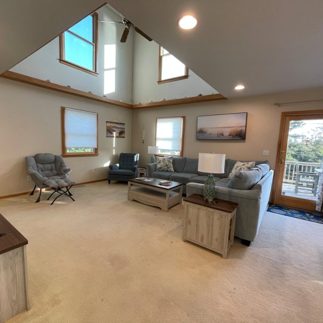 Living room with gray sectional, armchairs, coffee table, TV, large windows, and glass doors leading to a deck.