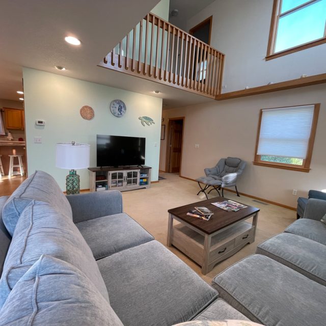 Living room with two gray sofas, TV, coffee table, armchair, and view into kitchen and upstairs hallway.
