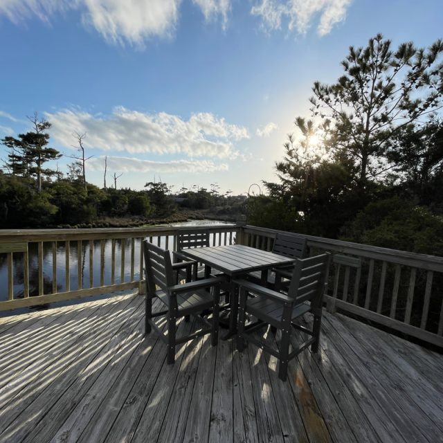 Wooden deck with a table and six chairs overlooks a calm river, surrounded by trees under a partly cloudy sky.