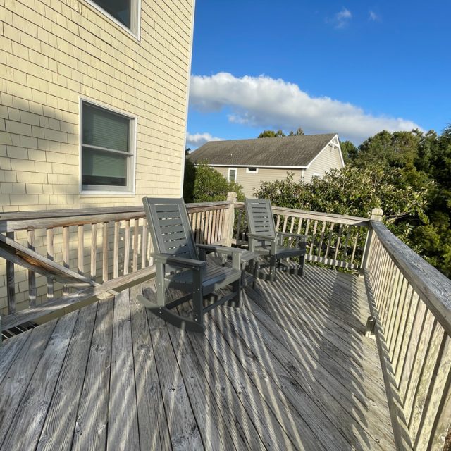 Two gray chairs on a weathered wooden deck with railing, attached to a house, trees and another house in background.