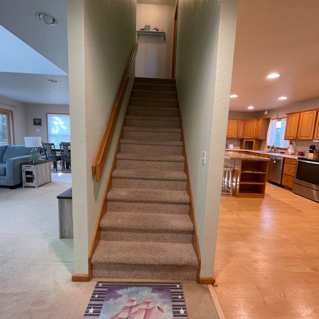 Carpeted stairs between a living room with blue sofas and a kitchen with wooden cabinets and light wood flooring.