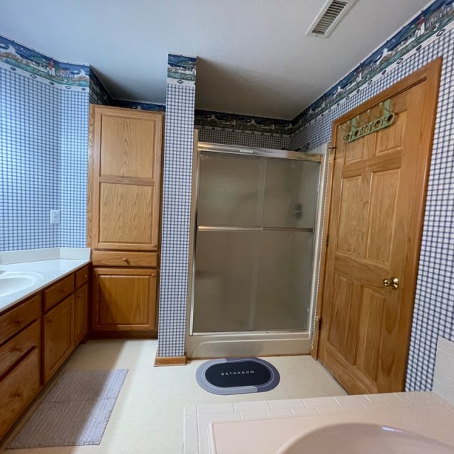 Bathroom with wood cabinets, glass shower, checkered wallpaper, double sinks, and a small rug on the floor.