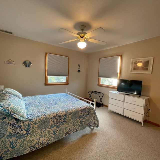 Bedroom with a queen bed, ceiling fan, two windows, dresser with TV, and beige walls.
