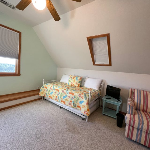 Bedroom with a daybed, colorful bedding, a striped chair, small TV on a nightstand, and two windows.