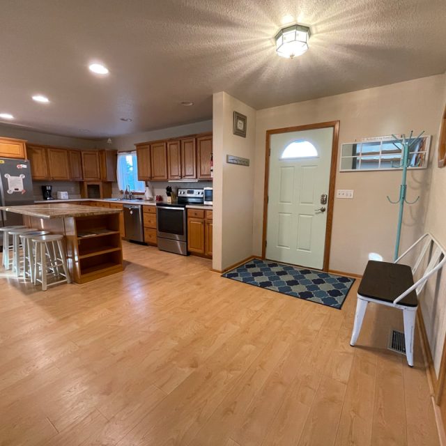 Spacious kitchen with wood cabinets, island, white bench, coat rack, and a door with a blue doormat.