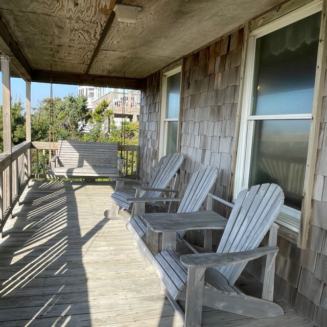 Wooden porch with two Adirondack chairs, a small table, and a porch swing facing trees and nearby houses.