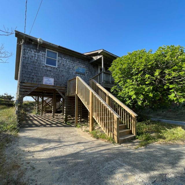 A wooden beach house on stilts with an exterior staircase, surrounded by greenery under a clear blue sky.