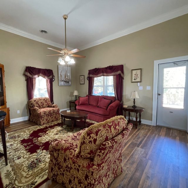 Living room with red patterned furniture, a rug, TV, bookshelves, and windows with red curtains; door is open.