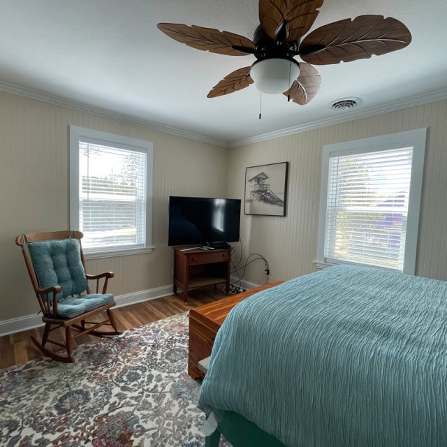 Bright bedroom with teal bedding, wooden rocking chair, TV on a stand, ceiling fan, and two windows with blinds.