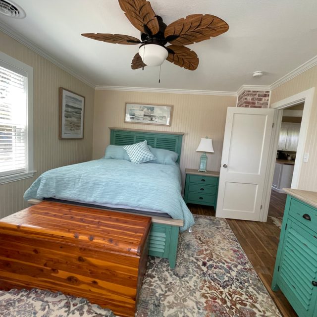 Bedroom with a teal bed, wooden chest, area rug, leaf-shaped ceiling fan, and natural light from a window.