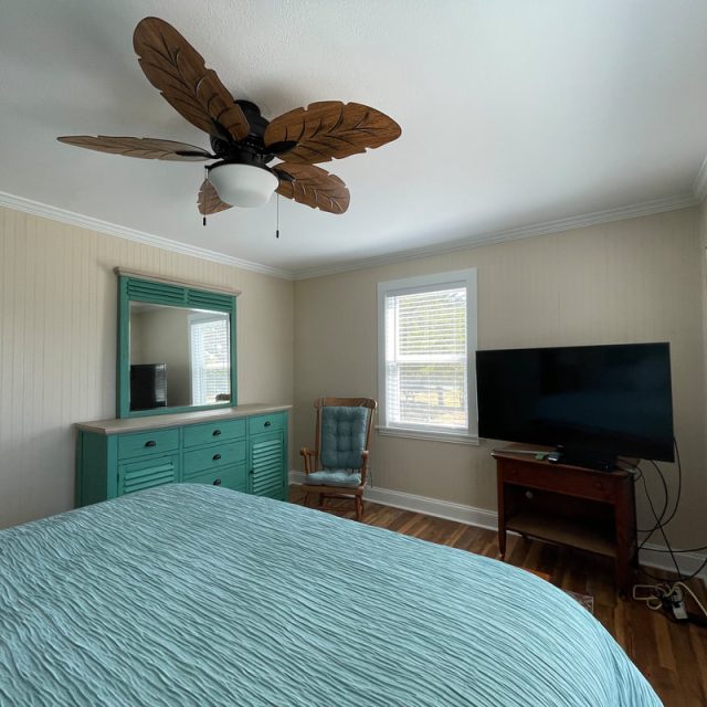 Bedroom with a leaf-shaped ceiling fan, teal dresser, rocking chair, bed, TV on a wooden table, and a window with blinds.