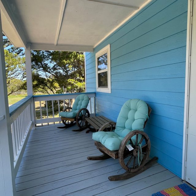 Two cushioned rocking chairs sit on a blue house porch with a view of trees and a road in the background.
