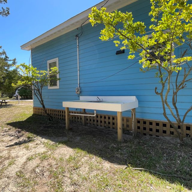 Outdoor sink with faucet stands next to a light blue house, surrounded by small trees and grass.