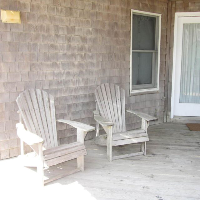 Two wooden Adirondack chairs sit on a weathered porch beside a window and a glass door with a doormat.