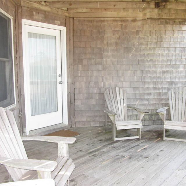 Wooden deck with three weathered Adirondack chairs, a glass door, and shingle siding under a covered porch.