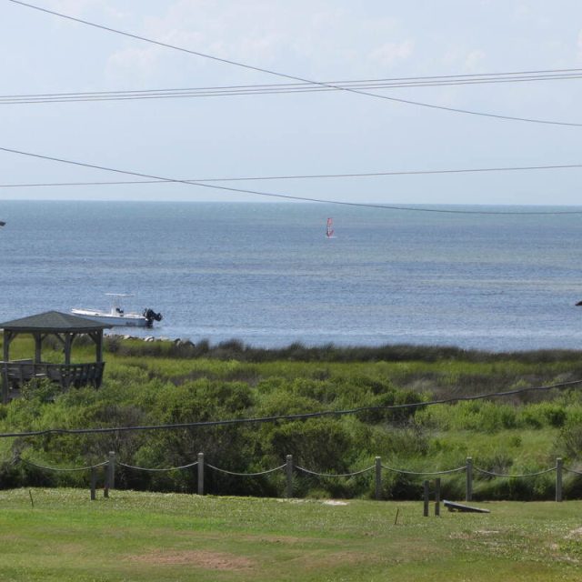 View of grassy dunes and buildings near the shore with a boat and a distant windsurfer on the calm sea under a clear sky.