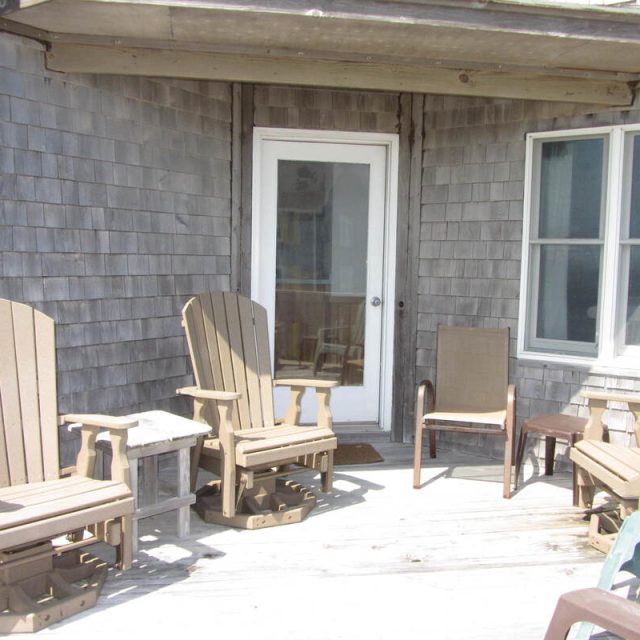 Four wooden chairs and a small table are arranged on a sunlit wooden deck outside a house with shingle siding.