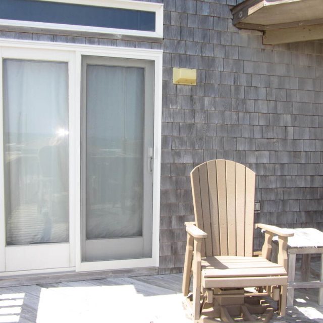 Two wooden chairs and a small table sit on a deck outside sliding glass doors on a shingled building.