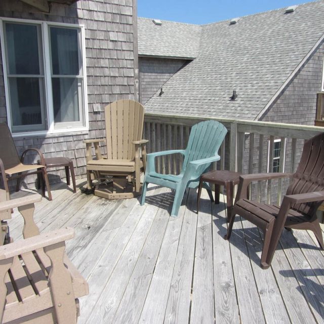 Wooden deck with several plastic and wooden chairs around the railing, next to a house with shingle siding.