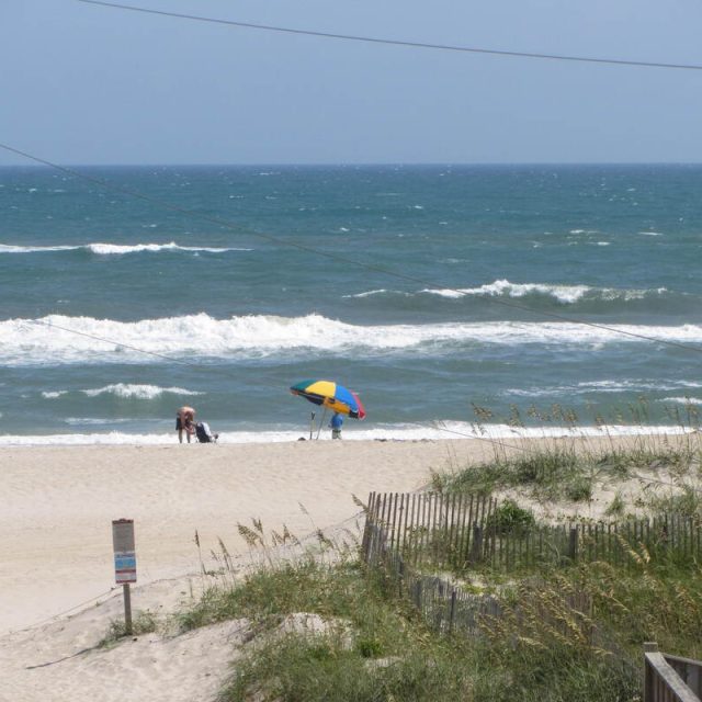 Two people with a colorful umbrella stand on a sandy beach near the ocean, with waves and sea grass visible.