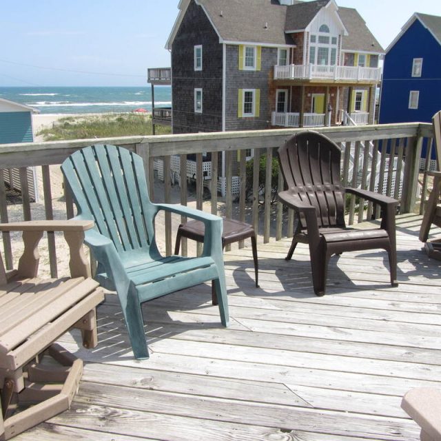 Several Adirondack chairs on a wooden deck overlook beach houses and the ocean under a partly cloudy sky.