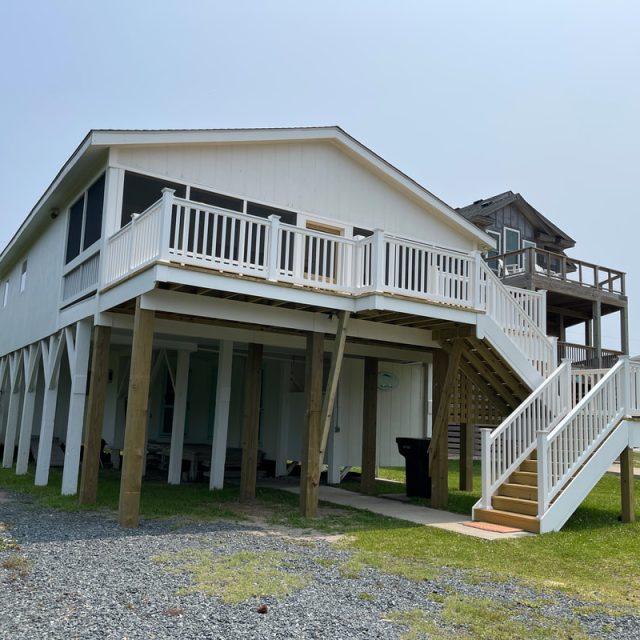 White elevated beach house with a large deck, wooden stairs, and gravel driveway on a sunny day.