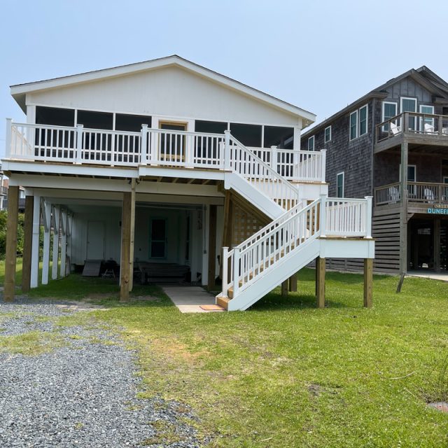 Two elevated beach houses with large porches and exterior stairs, set on grassy lots with gravel driveway.