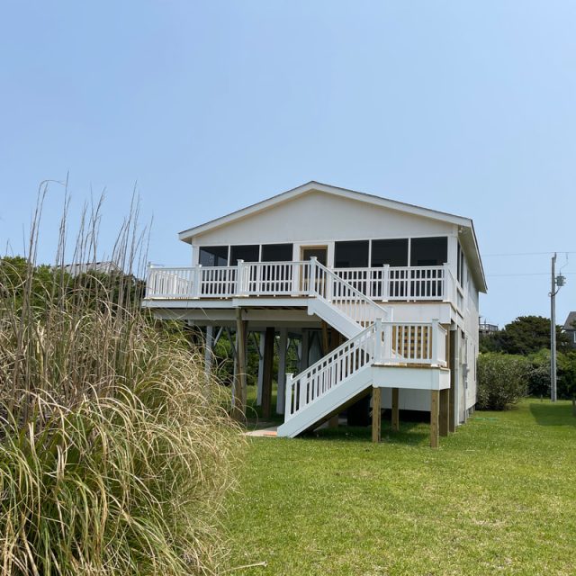White elevated beach house with a front staircase, screened porch, and grassy yard under a clear sky.