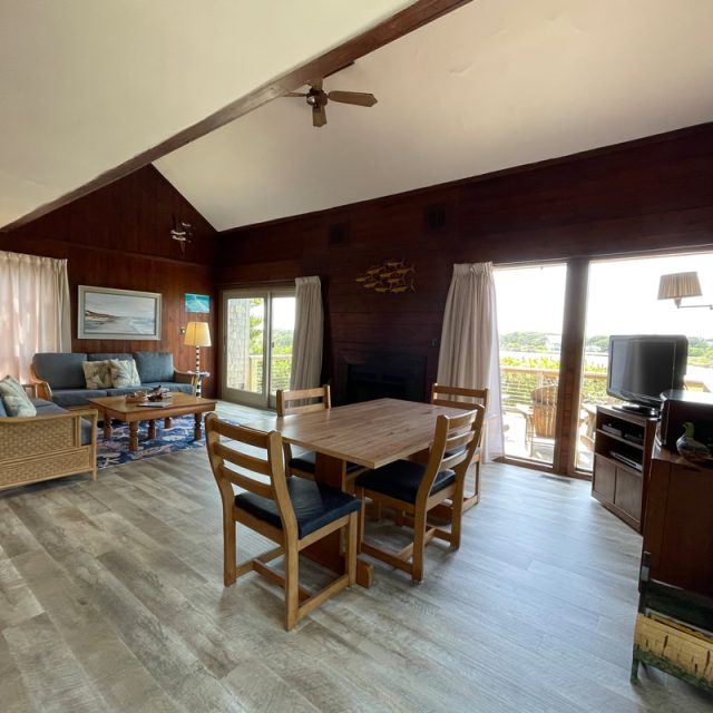Wood-paneled living room with dining table, sofas, TV, and large windows overlooking a deck and outdoor view.