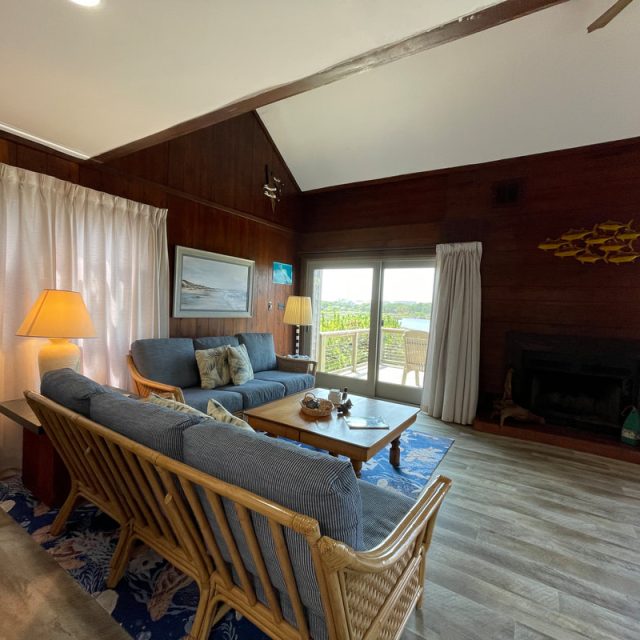 Living room with wood walls, rattan furniture, lamps, fireplace, and large window overlooking a balcony with outdoor view.