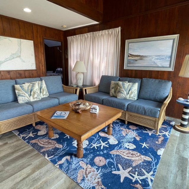 Living room with two wicker sofas, a wood coffee table, lamps, ocean-themed rug, and coastal artwork on wood-paneled walls.