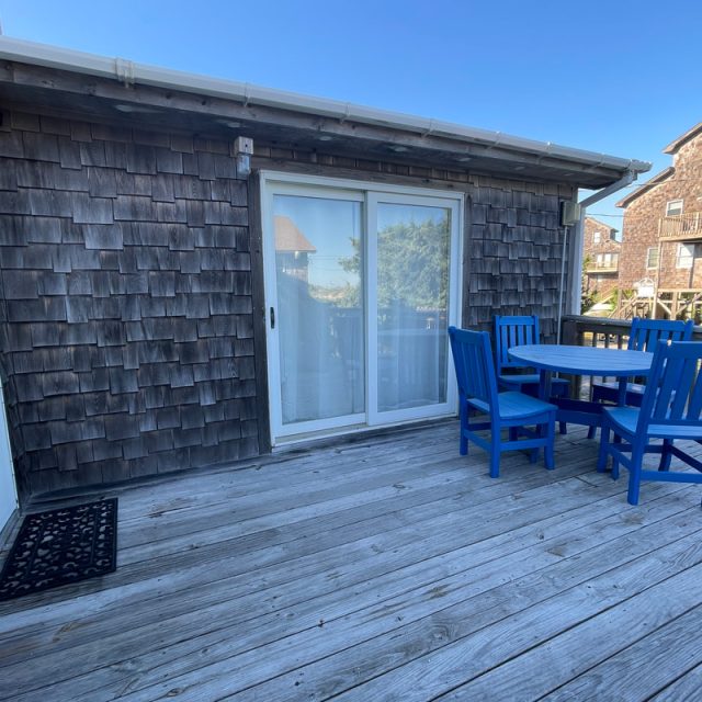 Wooden deck with a round table and four blue chairs, next to a building with weathered shingles and glass sliding doors.