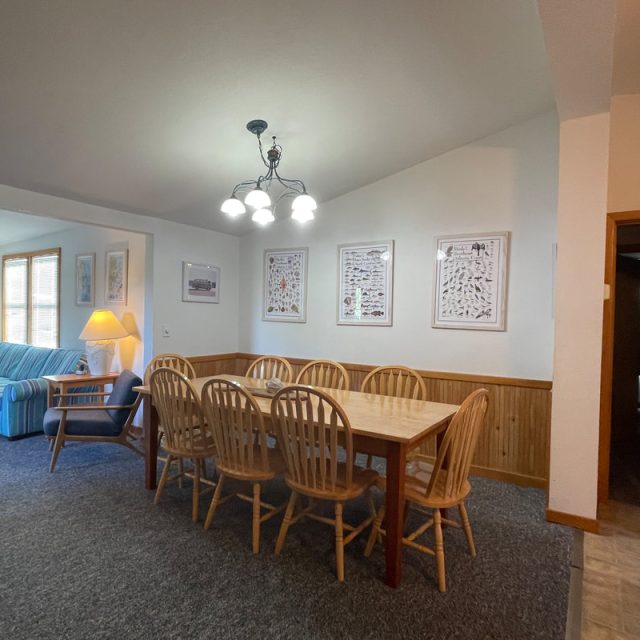 Dining area with a wooden table and eight chairs, adjacent to a living room with sofas and framed art on the walls.