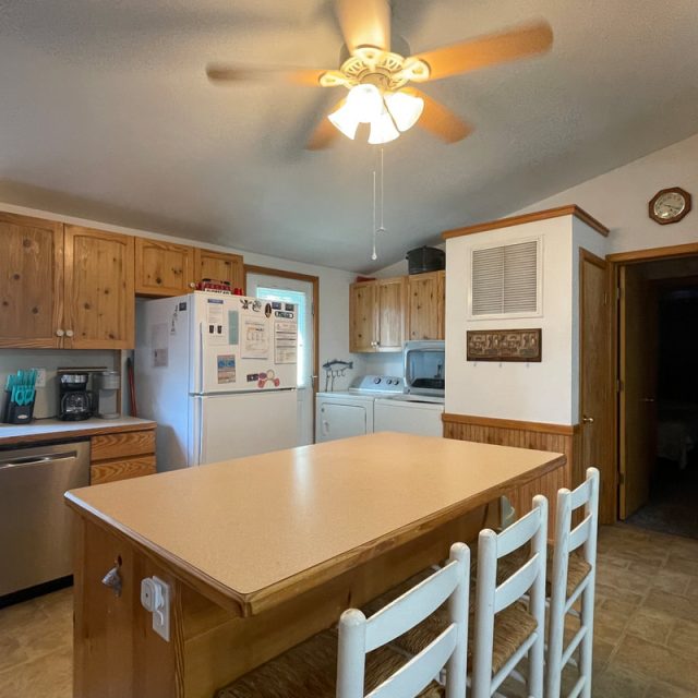 Kitchen with wooden cabinets, white appliances, central island, three chairs, ceiling fan, and wall clock.