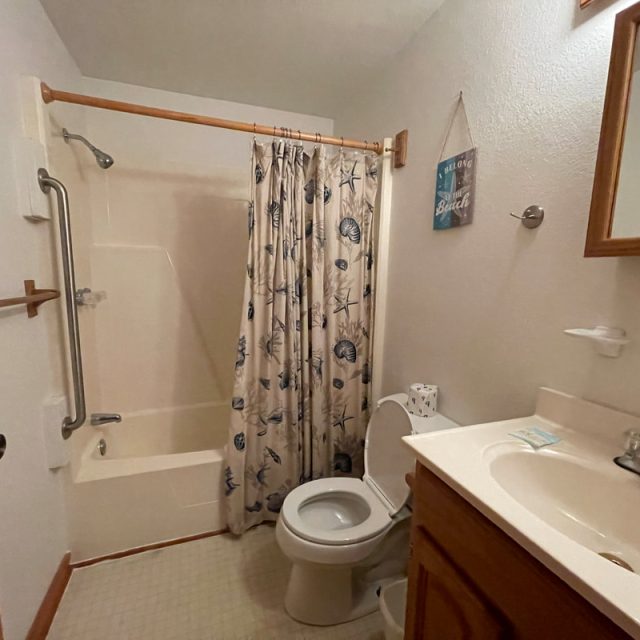 Small bathroom with a shower-tub combo, curtain with sea motifs, toilet, sink, and wooden accents.