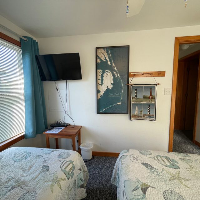 Small bedroom with two twin beds, wall art, a mounted TV, and sunlight streaming through a window with blue curtains.