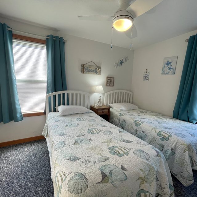 Bedroom with two twin beds, beach-themed bedding, blue curtains, a ceiling fan, and a lamp on a nightstand.