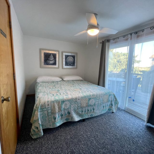 Small bedroom with a double bed, patterned quilt, ceiling fan, two wall art pieces, and sliding glass doors to a balcony.