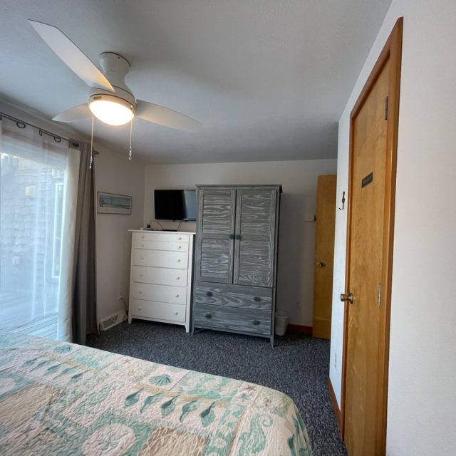 Bedroom with a bed, ceiling fan, dresser, armoire, small TV, and sliding glass door with sheer curtains.