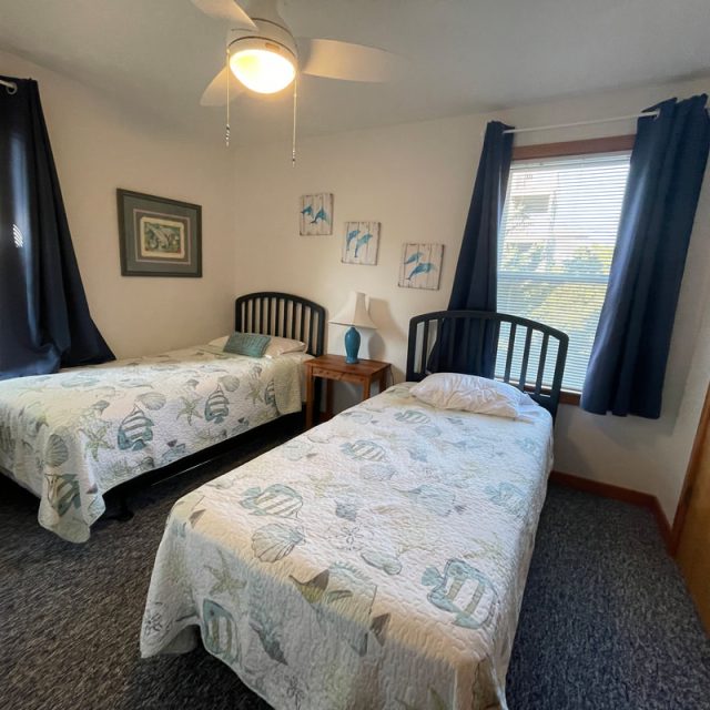 Bedroom with two twin beds, blue curtains, wall art, a nightstand, lamp, ceiling fan, and windows with natural light.