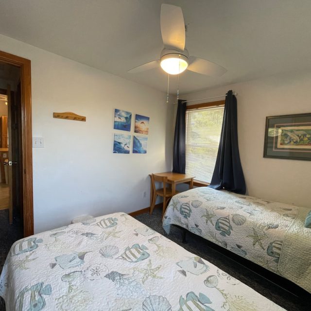 Twin beds with seashell quilts in a small, bright bedroom with a ceiling fan, window, and a wooden desk by the window.
