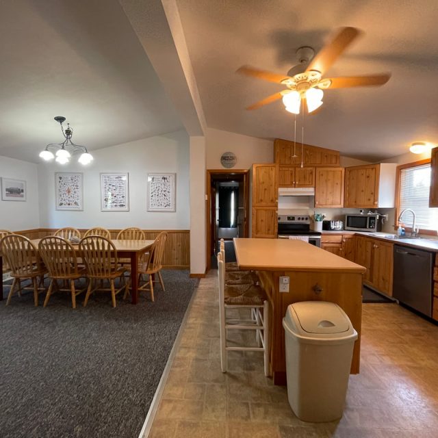 Open kitchen and dining area with wooden cabinets, large dining table, ceiling fans, and natural lighting.