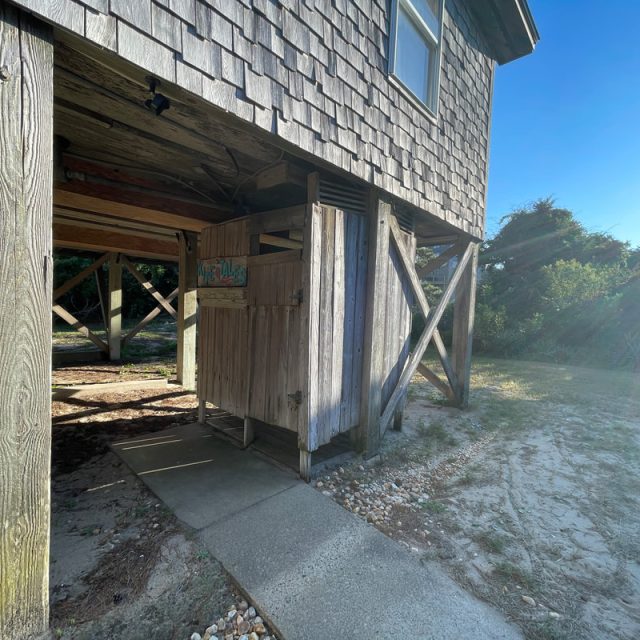 Wooden outdoor shower under a raised house, with sunlight streaming from the right and a concrete path leading to it.
