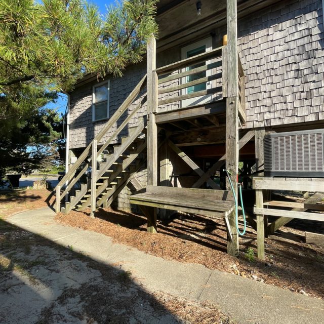 Wooden stairs lead to a raised house with shingle siding and an air conditioning unit, surrounded by trees and sunlight.