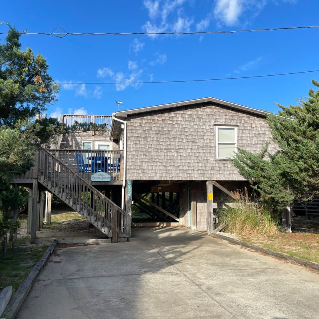 Wooden beach house on stilts with exterior stairs, surrounded by trees, under a clear blue sky.