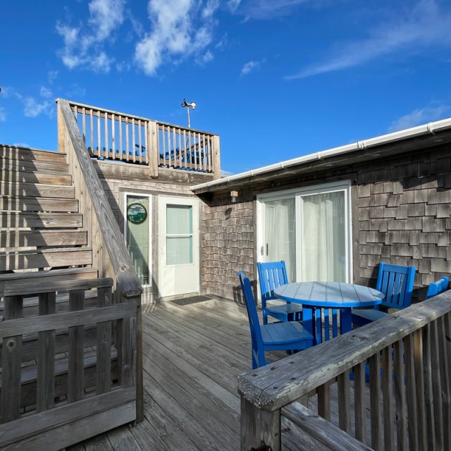 Wooden deck with blue outdoor dining set, stairs leading to an upper level, and sliding glass door under a blue sky.
