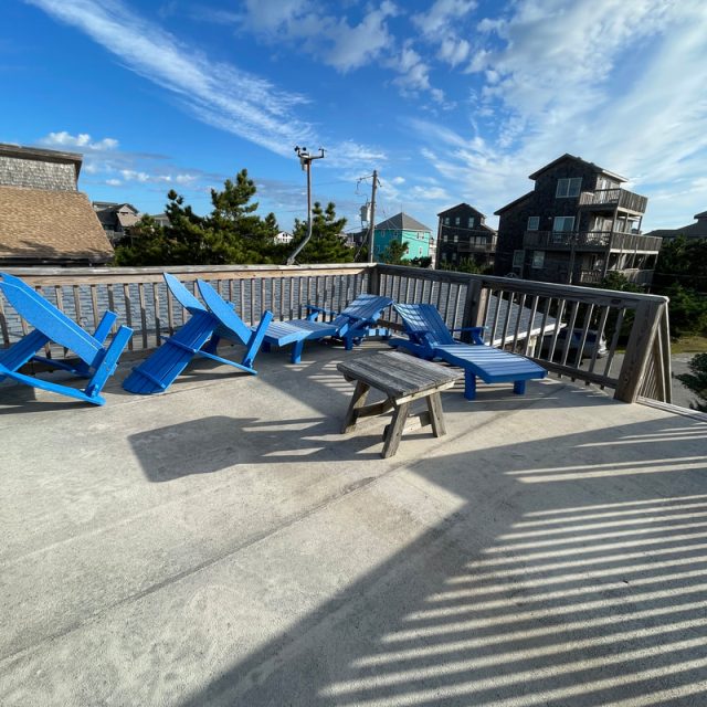 Five blue lounge chairs and a wooden stool on a sunny rooftop deck, surrounded by houses and trees under a blue sky.