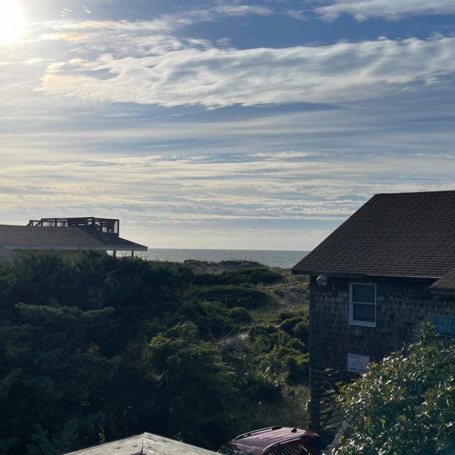 Sunlit sky over coastal houses and greenery with the ocean visible in the background under scattered clouds.